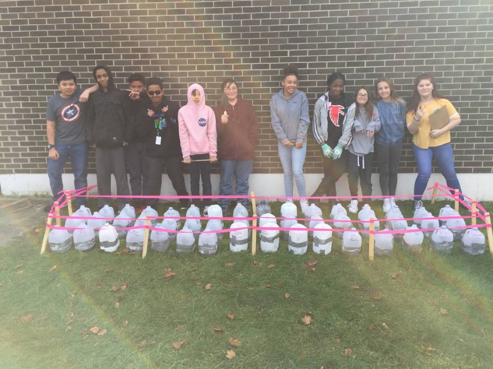 Students stand behind milk jugs filled with soil and native plant seeds